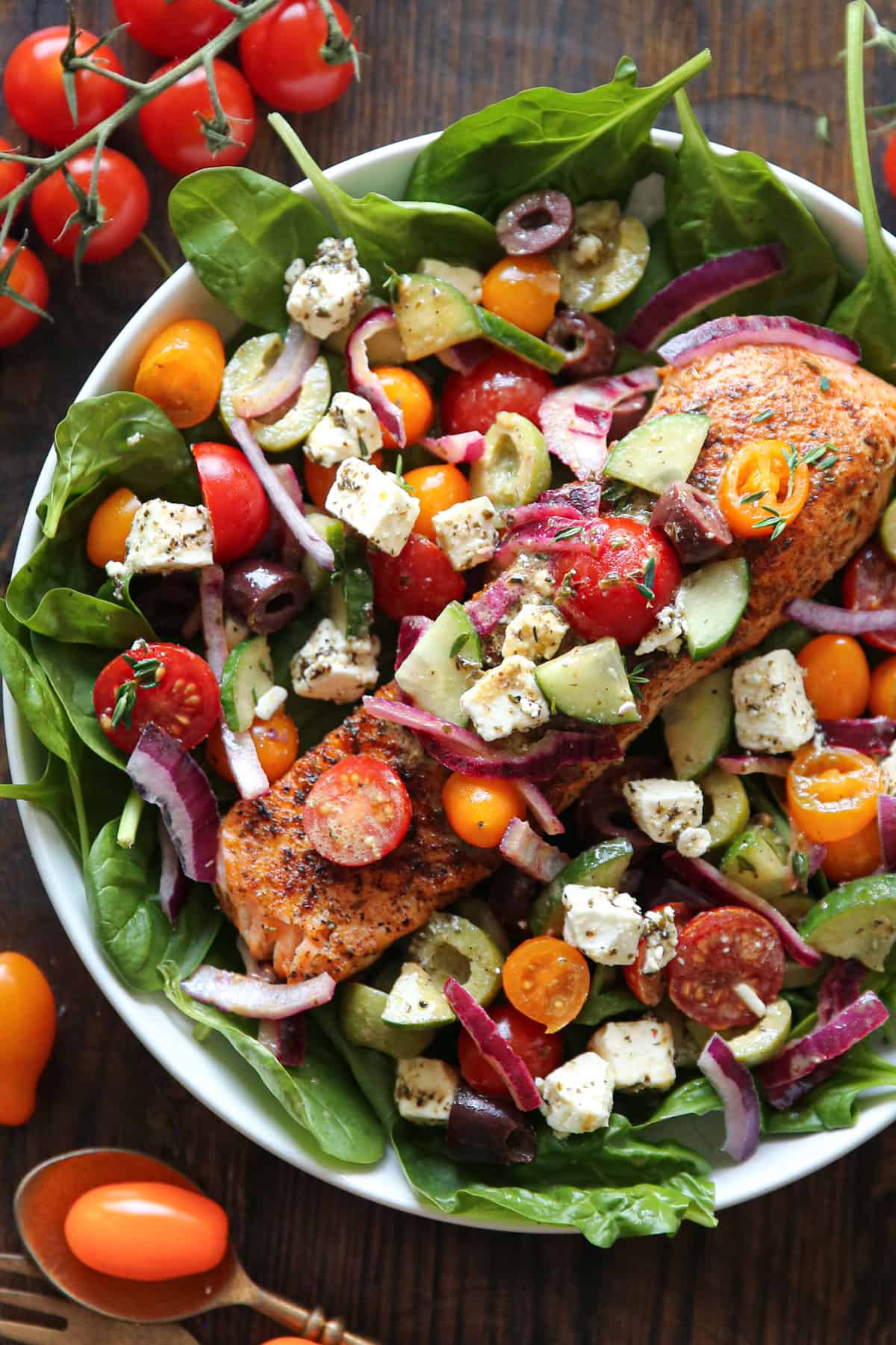 Greek Salmon Salad (with spinach, kalamata olives, green olives, cherry tomatoes, cucumber, red onion, and tangy feta) in a white bowl.