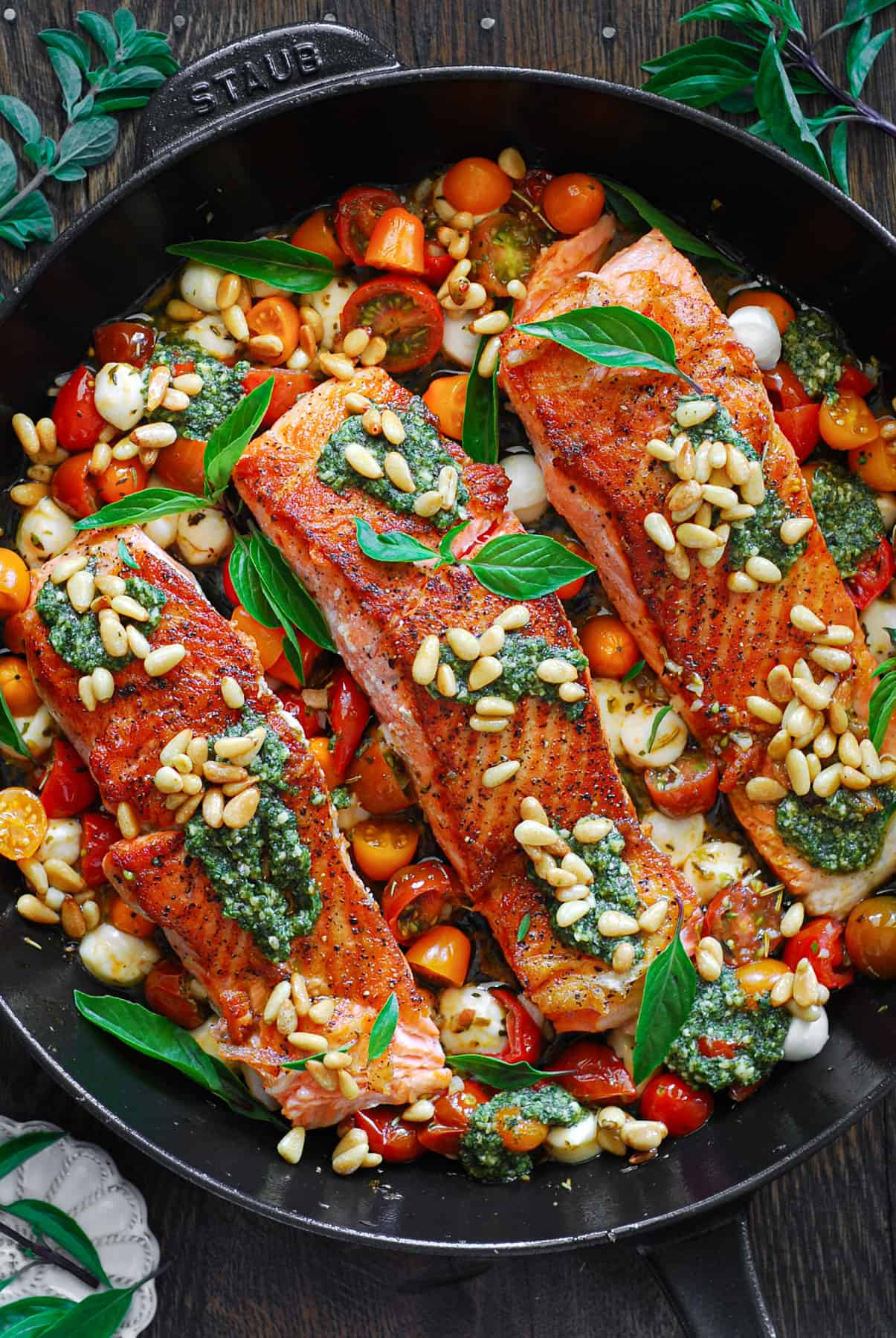  Pesto Salmon with cherry or grape tomatoes, Mozzarella cheese, basil pesto (​homemade​ or store-bought), garlic, lemon, oregano, and toasted pine nuts in a cast iron skillet.