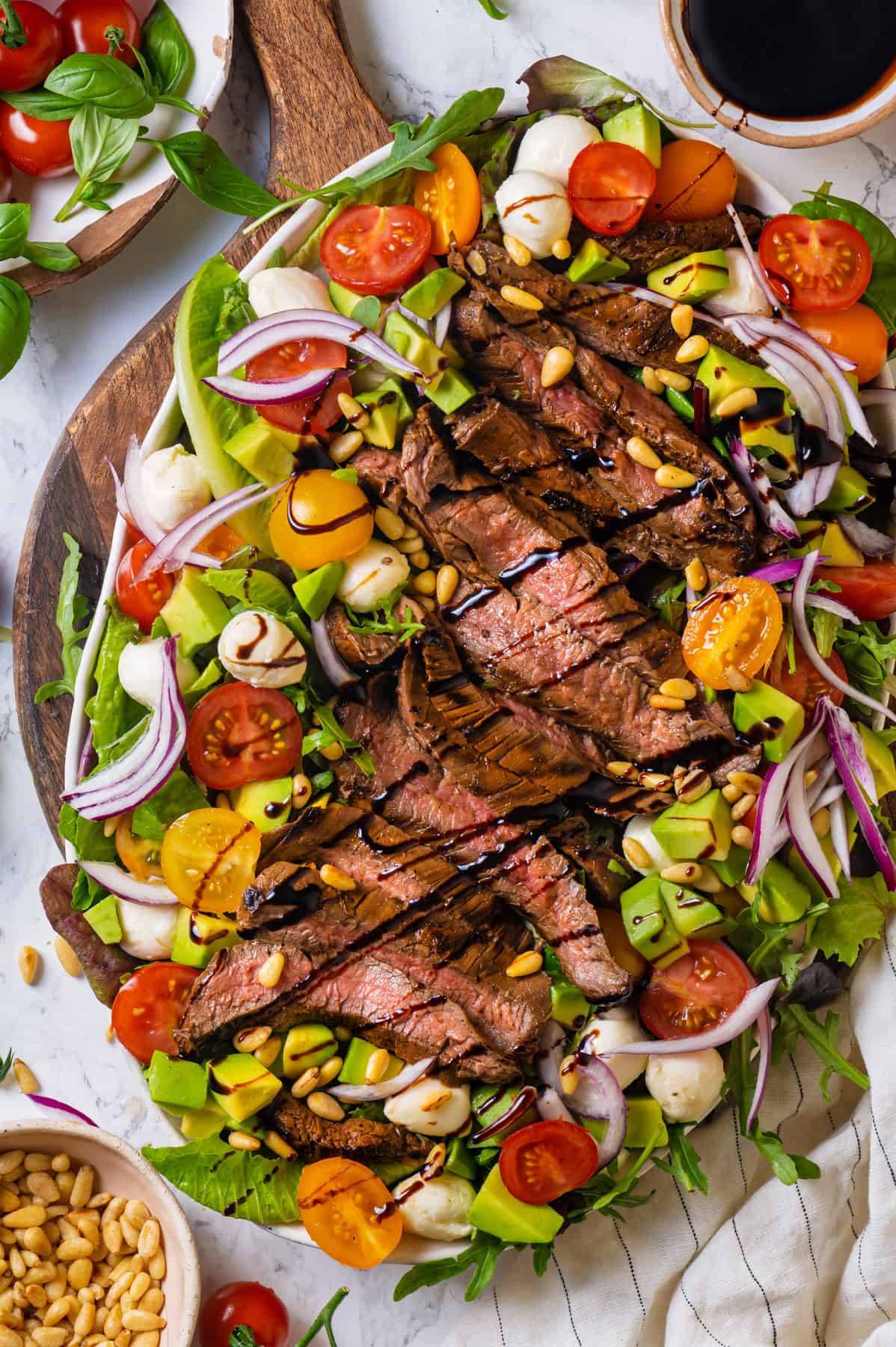 Grilled (Marinated) Flank Steak Salad (with baby greens, arugula, cherry tomatoes, red onions, mozzarella pearls, avocado, pine nuts, drizzled with balsamic glaze) on a white platter.
