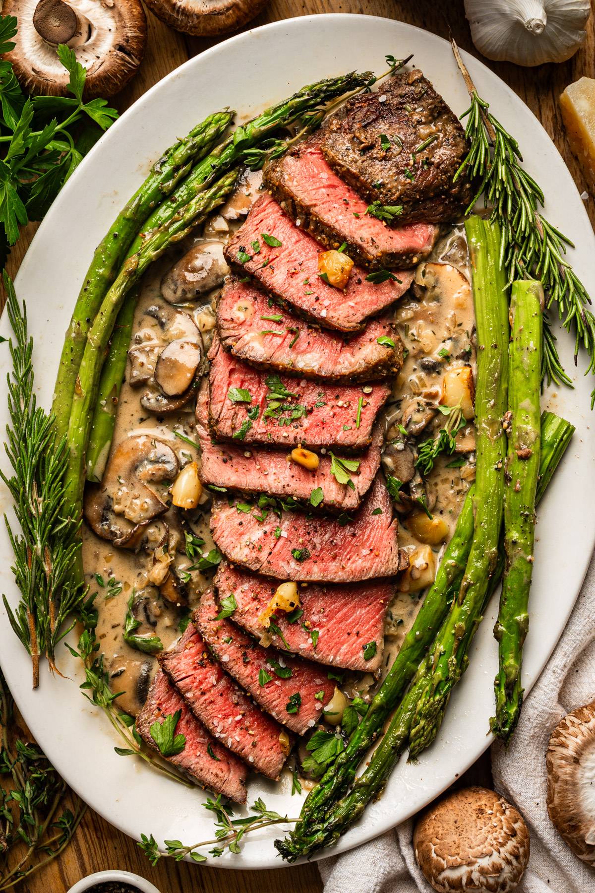 Sliced pan-seared steak with mushroom sauce and asparagus on a white elongated platter.