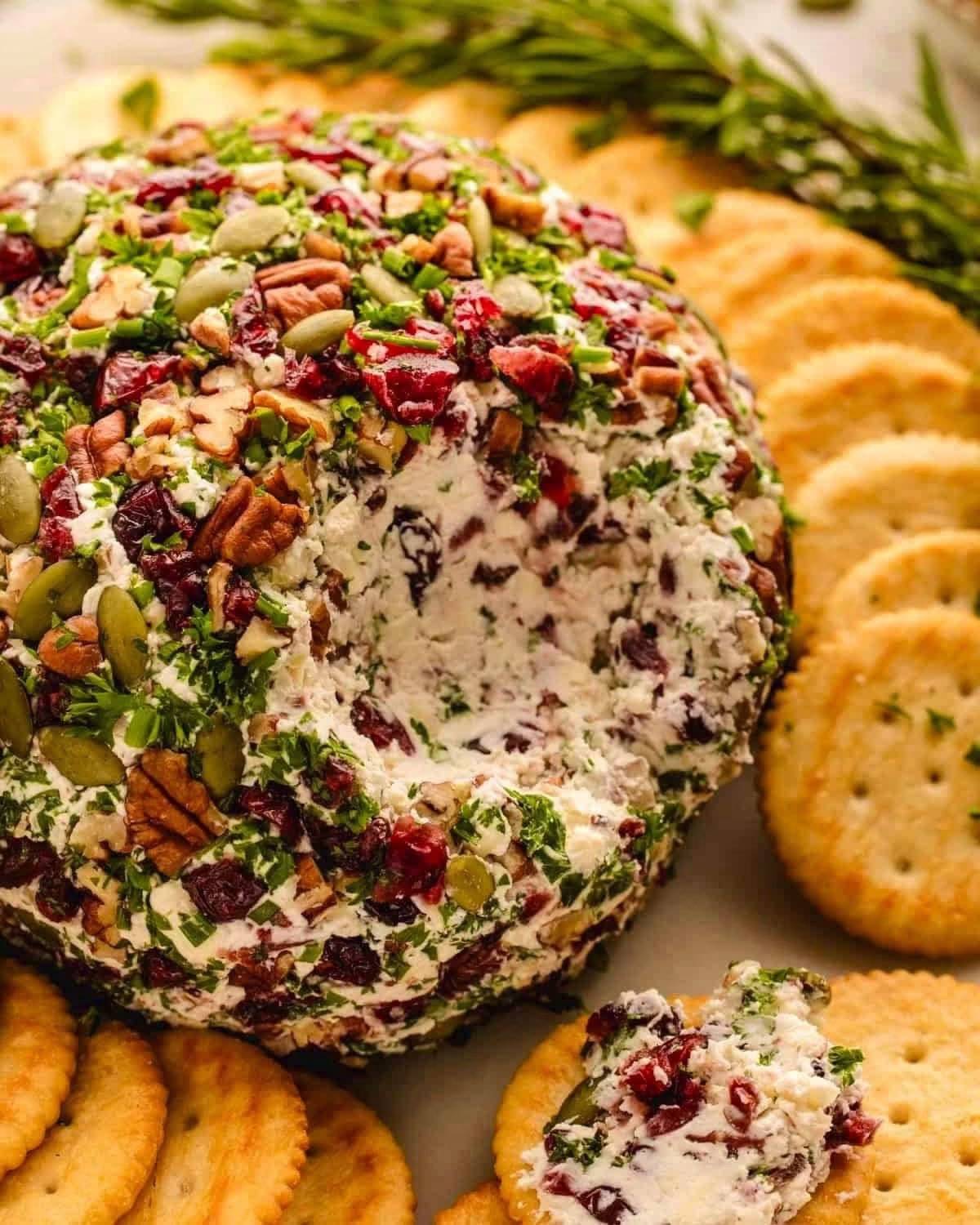 Cheese Ball covered with dried cranberries, pecans, and pepitas, surrounded by crackers on a white plate.