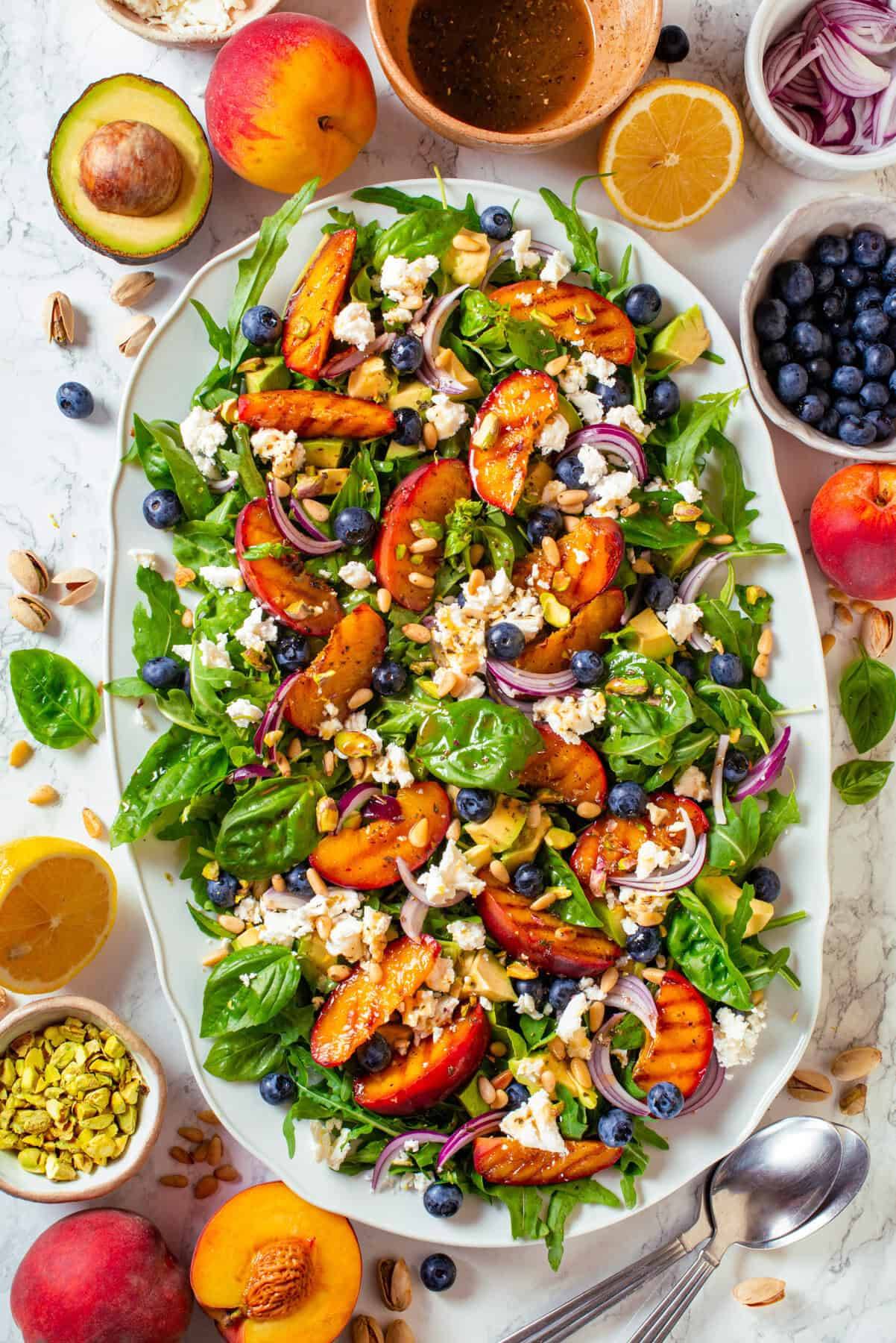 Grilled peach salad with arugula, avocado, berries, toasted nuts on a white platter.