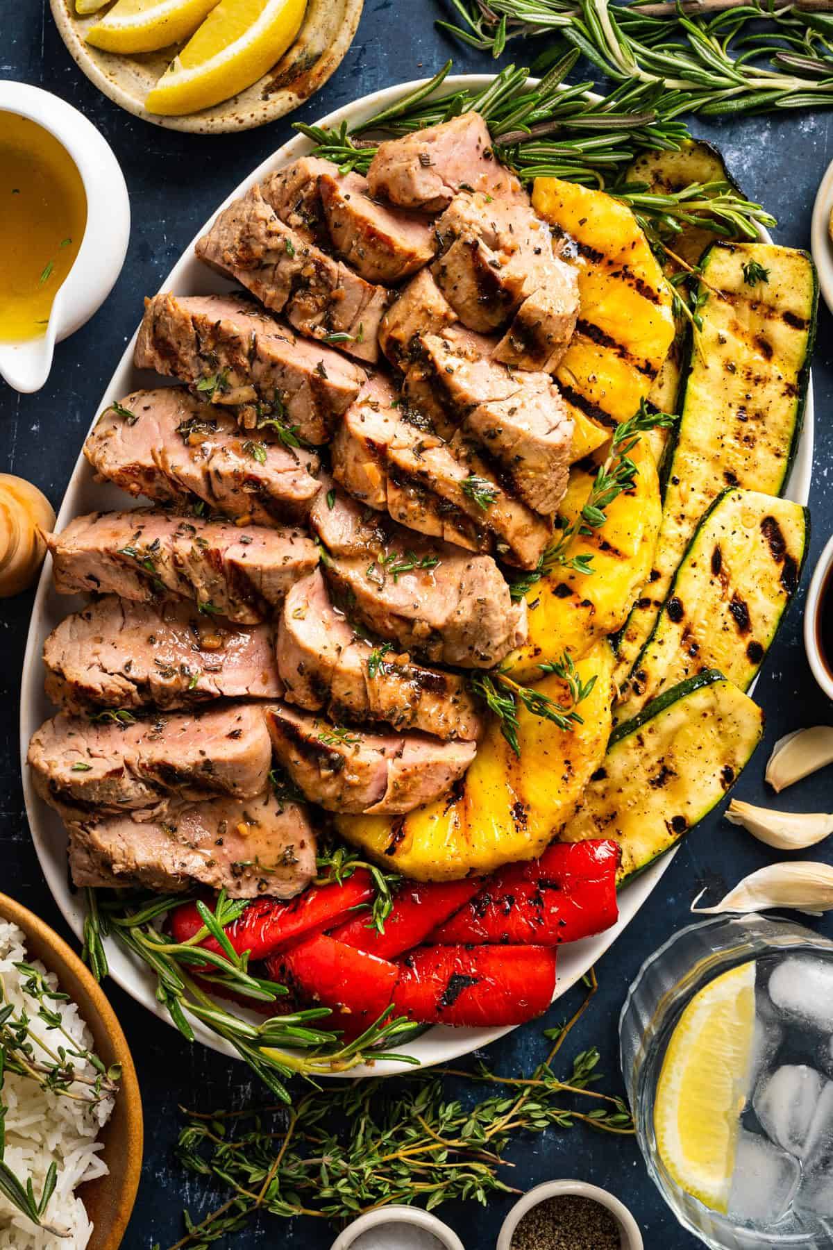 Marinated Grilled Pork Tenderloin with Caramelized Pineapple, Grilled Veggies, and Garlic Herb Butter on a white platter.