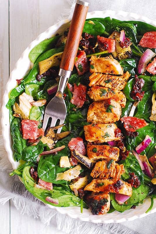 Italian Salad with Chicken, Spinach, Artichokes, Salami, Sun-Dried Tomatoes, Red Onions in a white bowl.