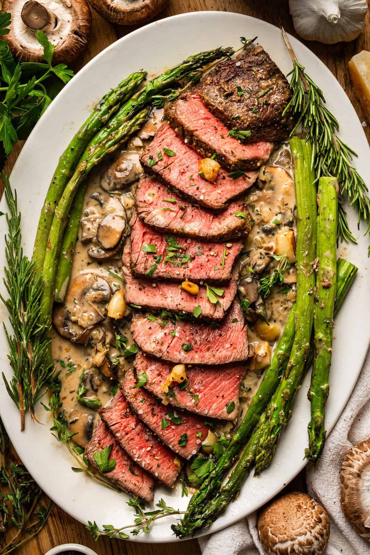 Pan-Seared Steak with Creamy Mushroom Sauce, Roasted Garlic, & Asparagus on a white platter.