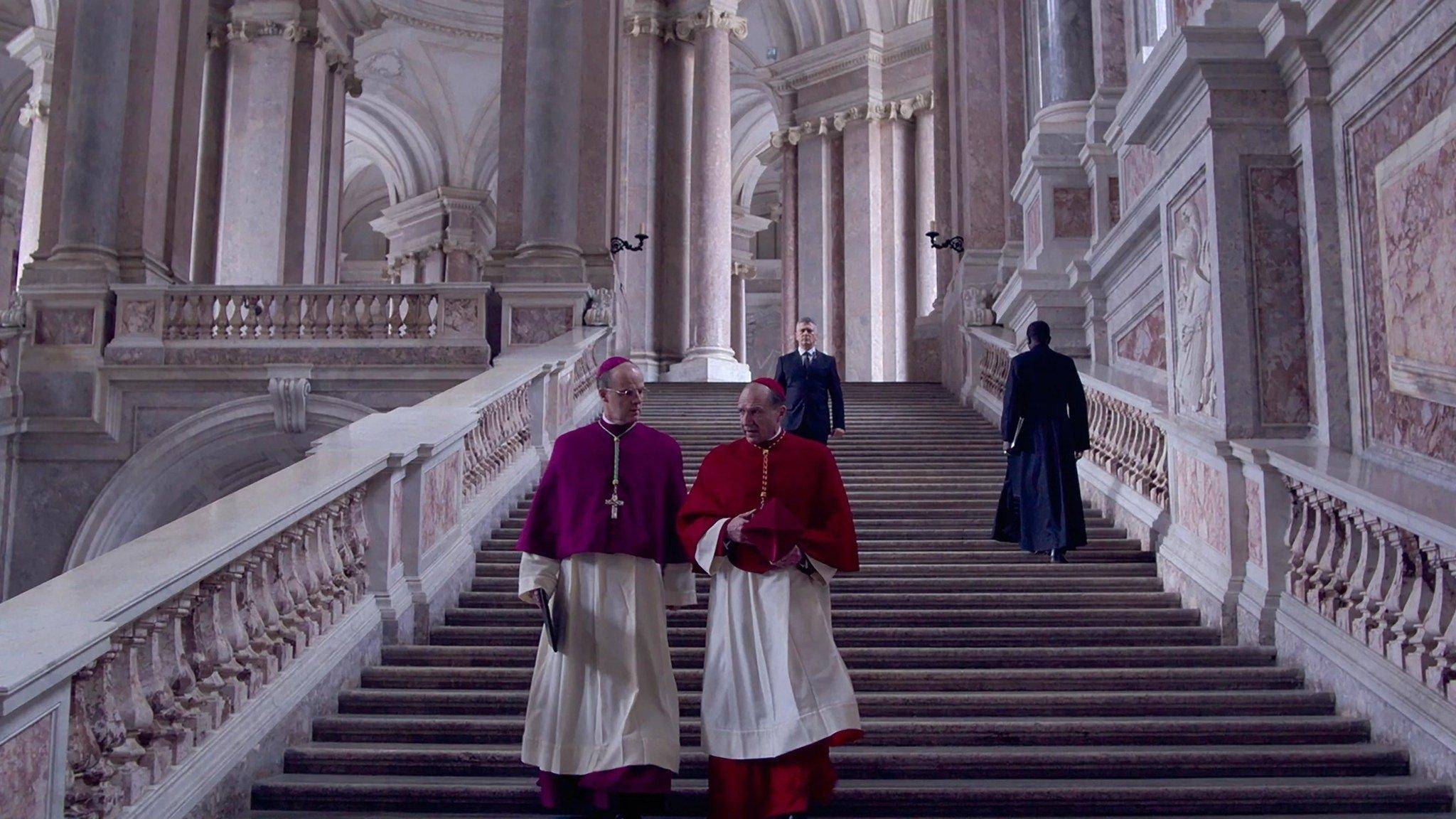 Two Catholic priests talk in serious tones as the descend a lavish staircase.