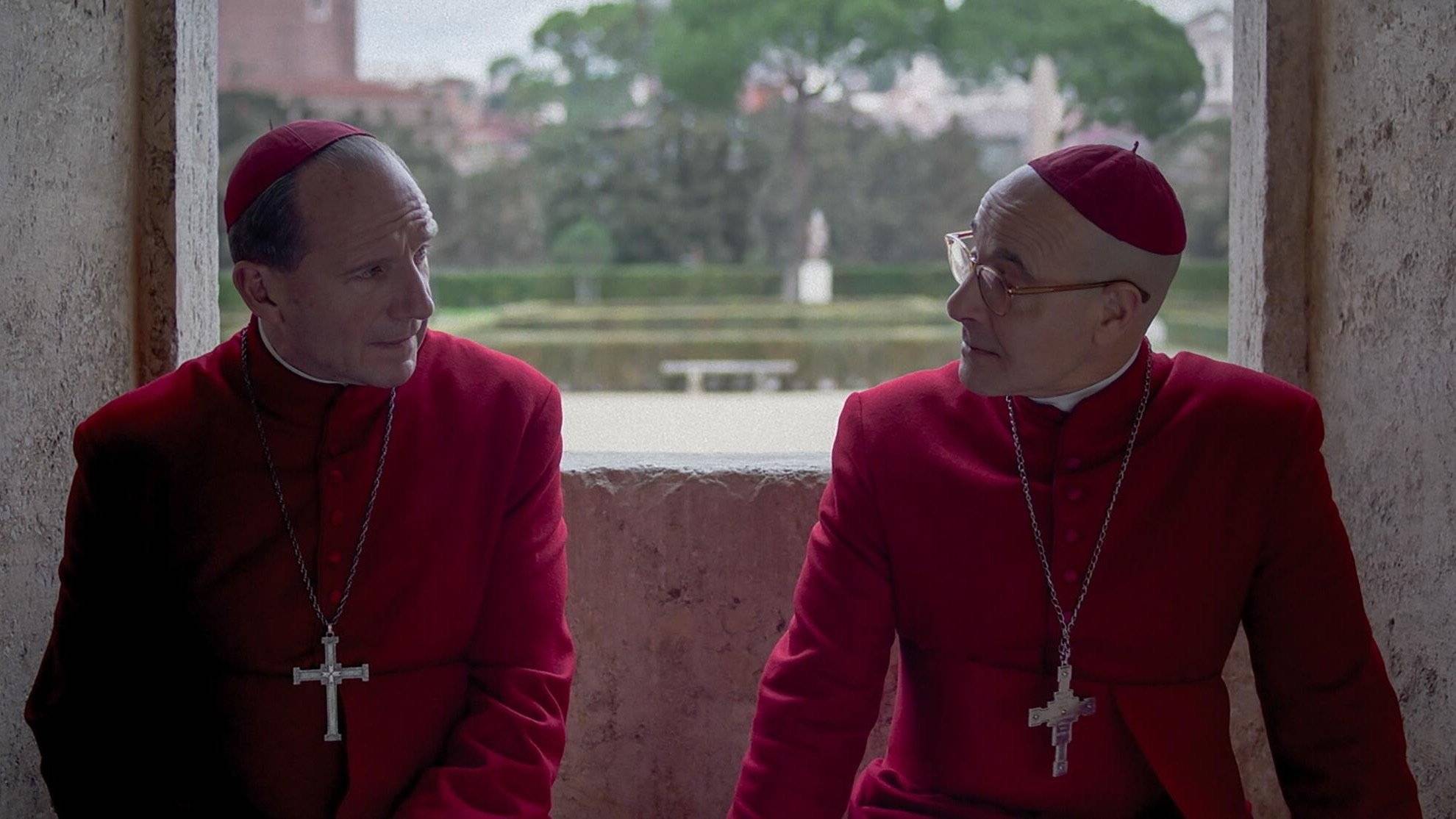 Two Catholic priests wearing red robes and crosses sit in deep discussion, they look tense.