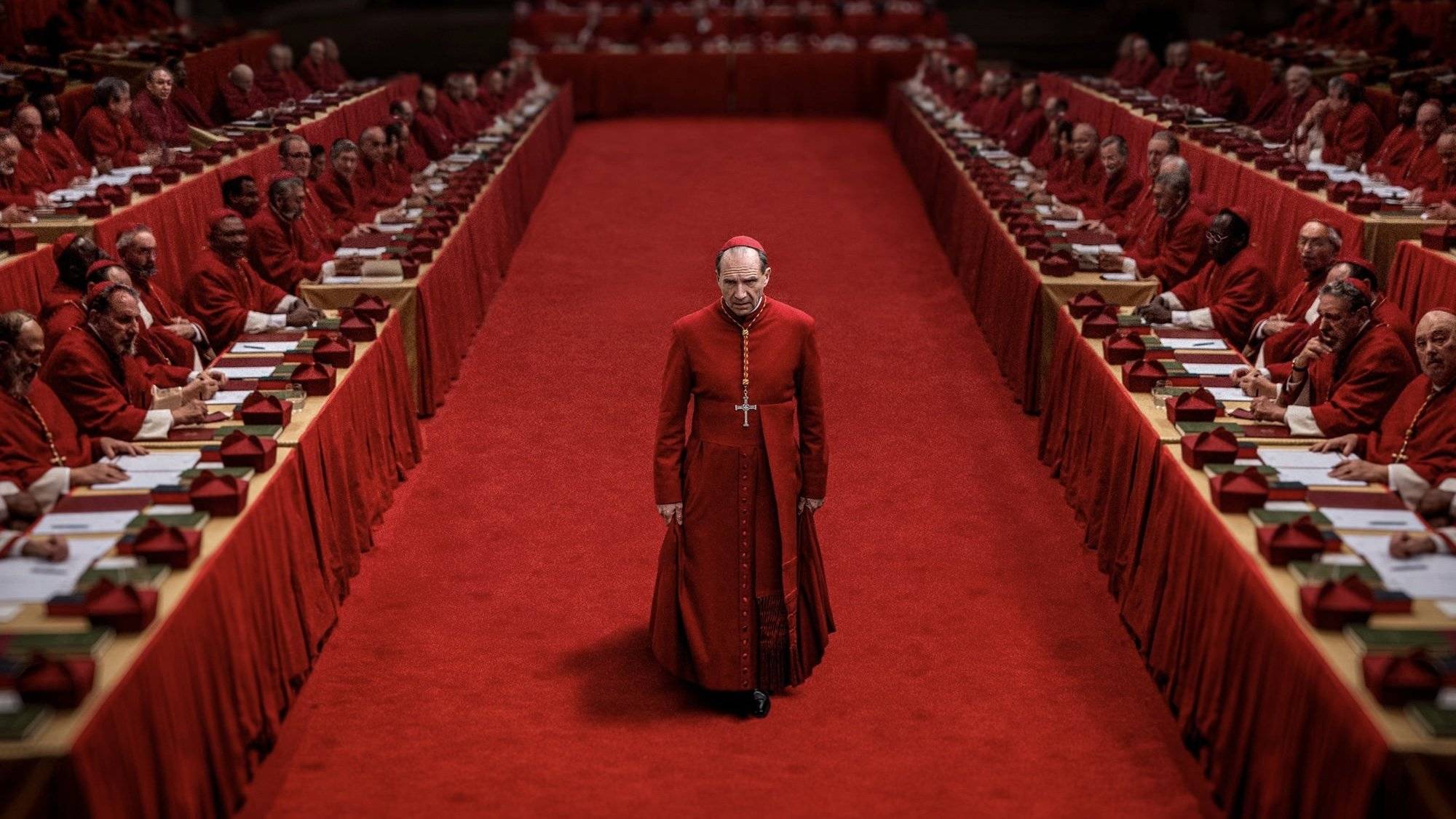 A Roman Catholic Cardinal stands on vivid red carpet among tables of seated priests.