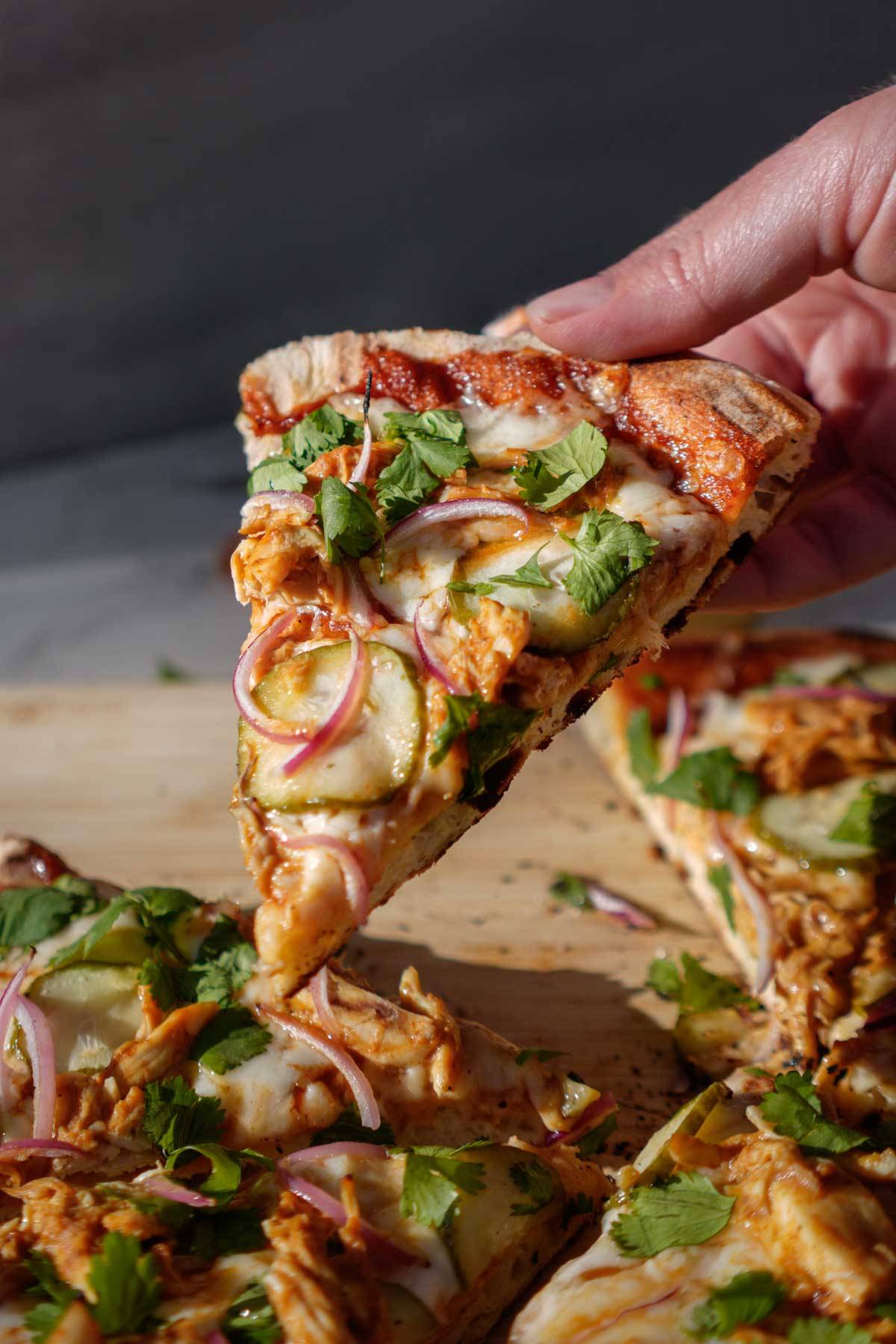 Picture of a hand holding up a slice of BBQ chicken and pickle pizza