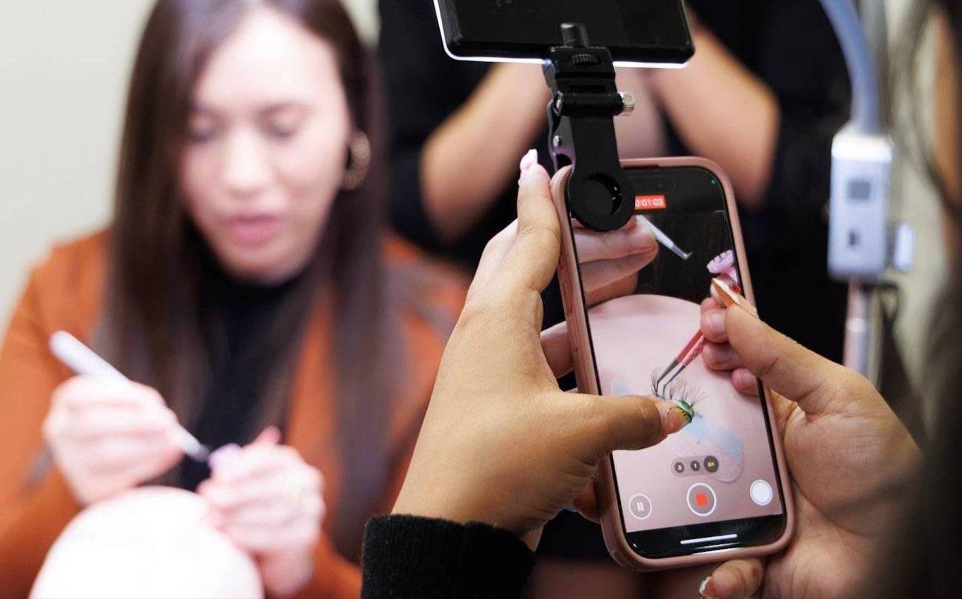 Lash artist applying lashes while a phone on a tripod films the process during a D'Lash training
