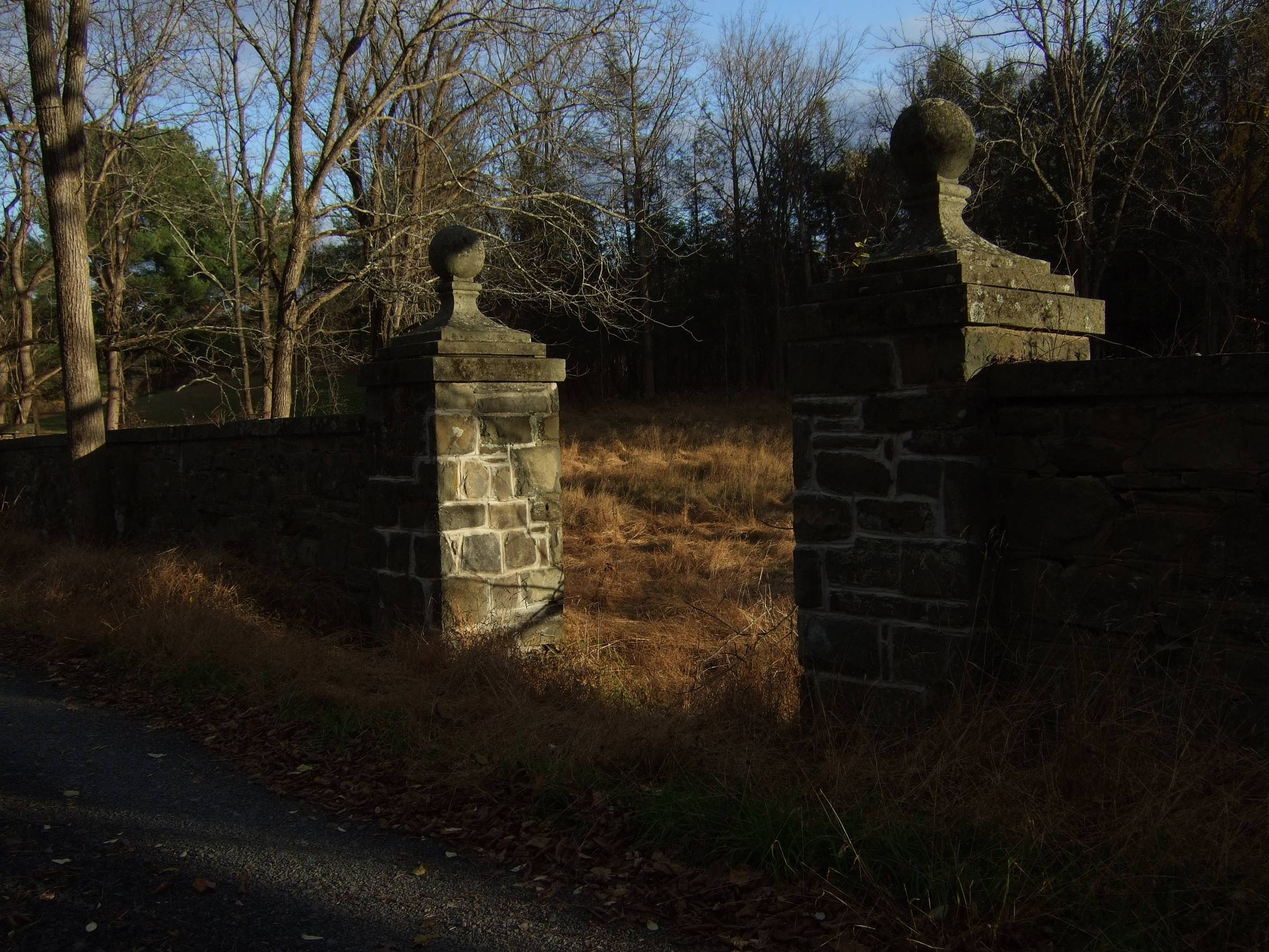 Weathered stone gateposts and a low stone wall stand at the edge of an overgrown field surrounded by bare trees. The house and estate the entrance once belonged to are no longer there.