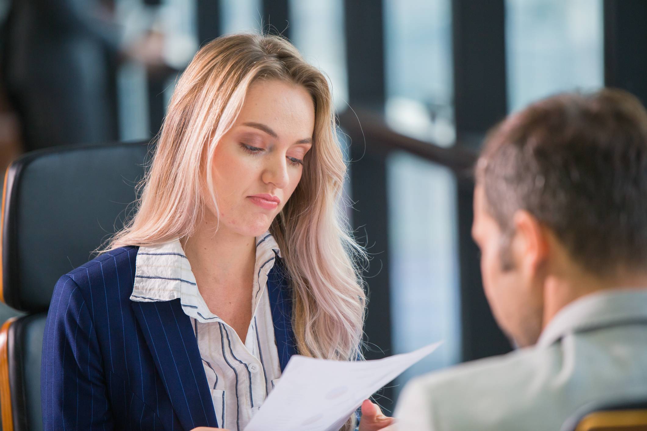 Blond woman in blue pin stripe blazer holds a paper and looks indifferent. Blurred in front of her is the back of a male in a lighter coloured suit.