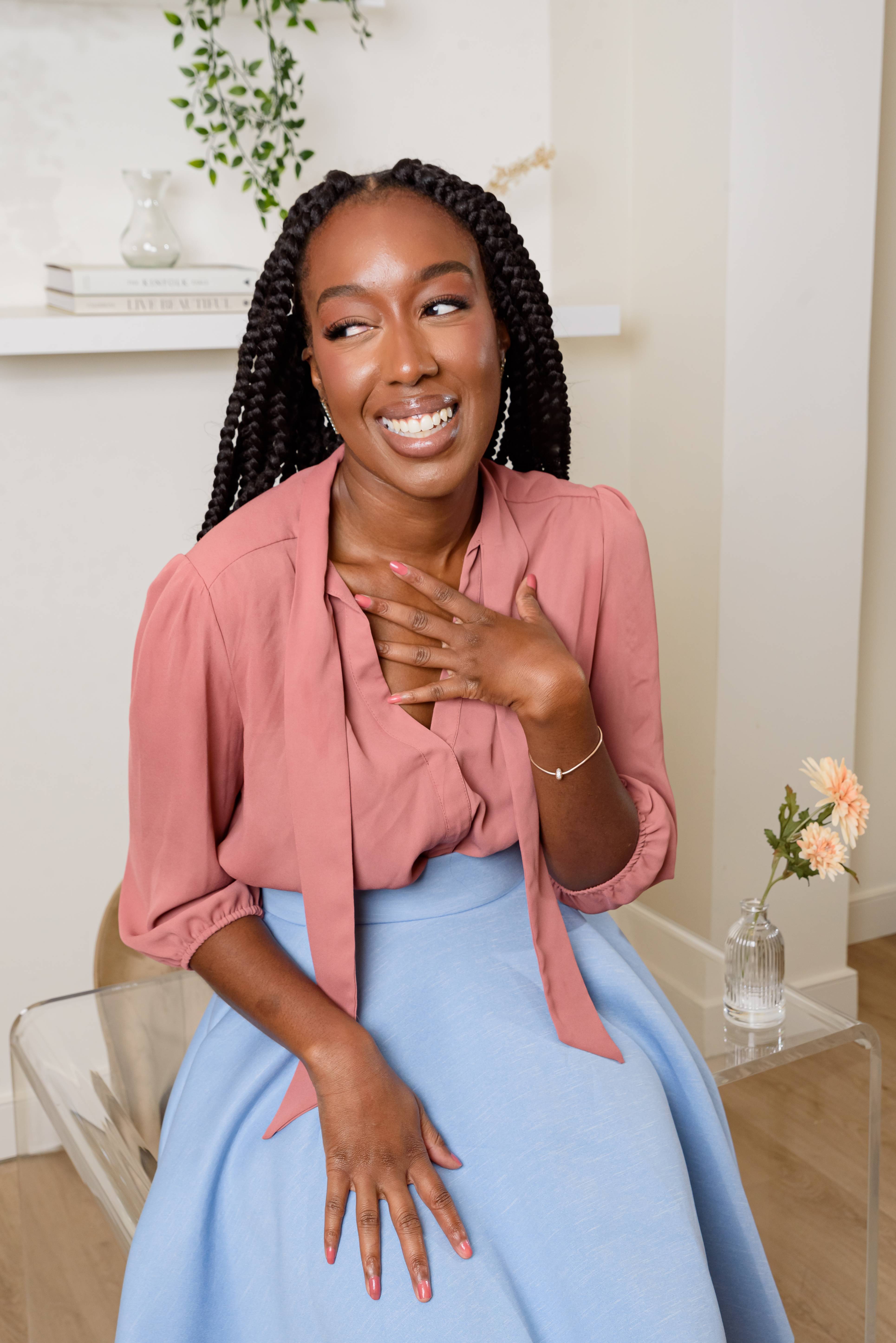 A woman with long braided hair is smiling while sitting in a room. She is wearing a pink blouse and a light blue skirt. One hand is on her chest and the other rests on her lap. In the background, there's a shelf with books and a potted plant, and a small vase with flowers on a clear table beside her.