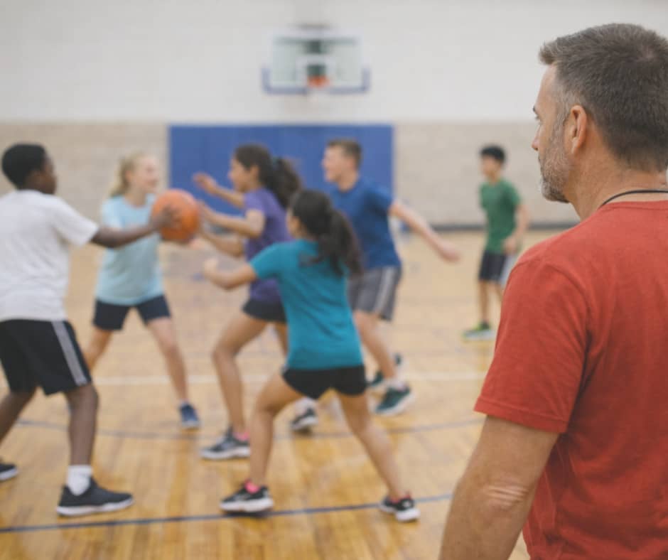 PE teacher observing students playing a small-sided basketball game in a gym