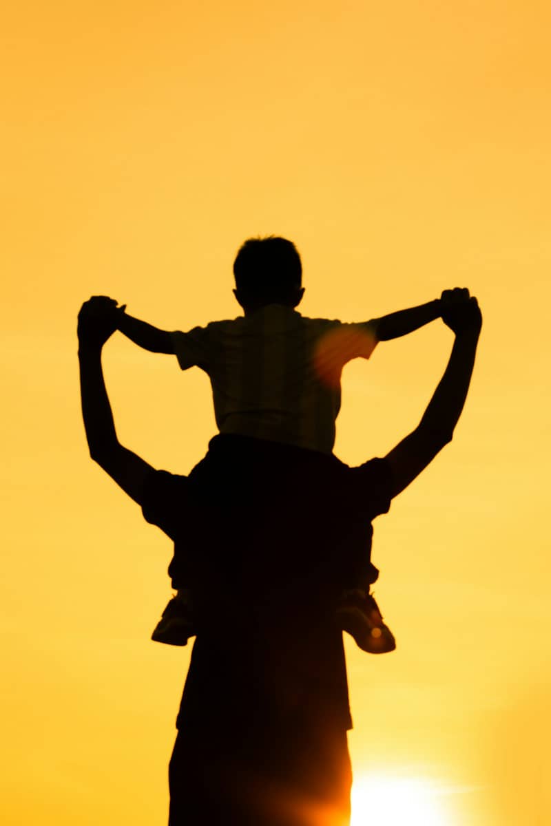 A child sitting on an adult's shoulders in silhouette against a warm orange sky