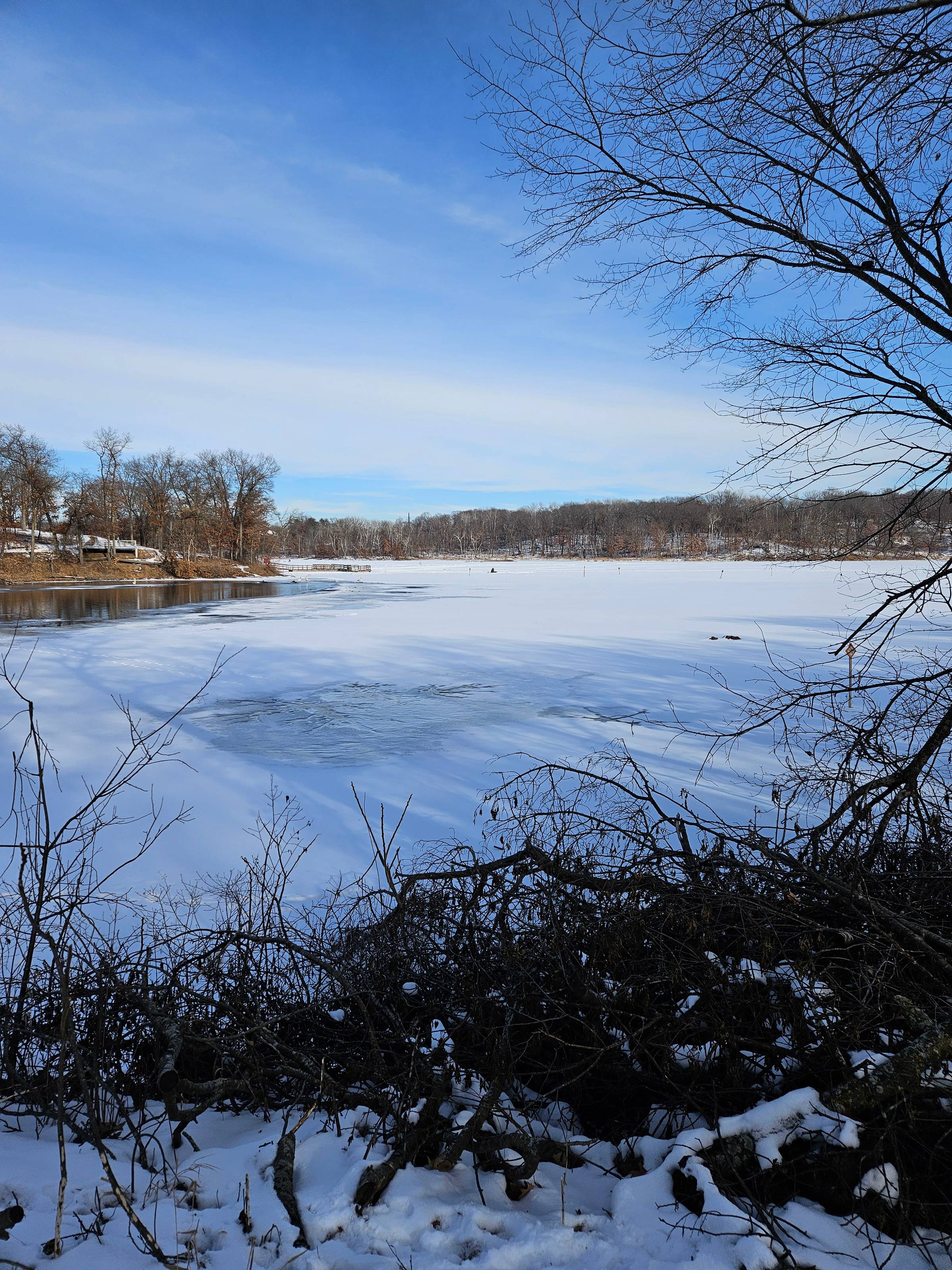 A photograph of an ice-covered lake, with bare trees on the shore and a blue sky that has some white streaks of clouds across it.