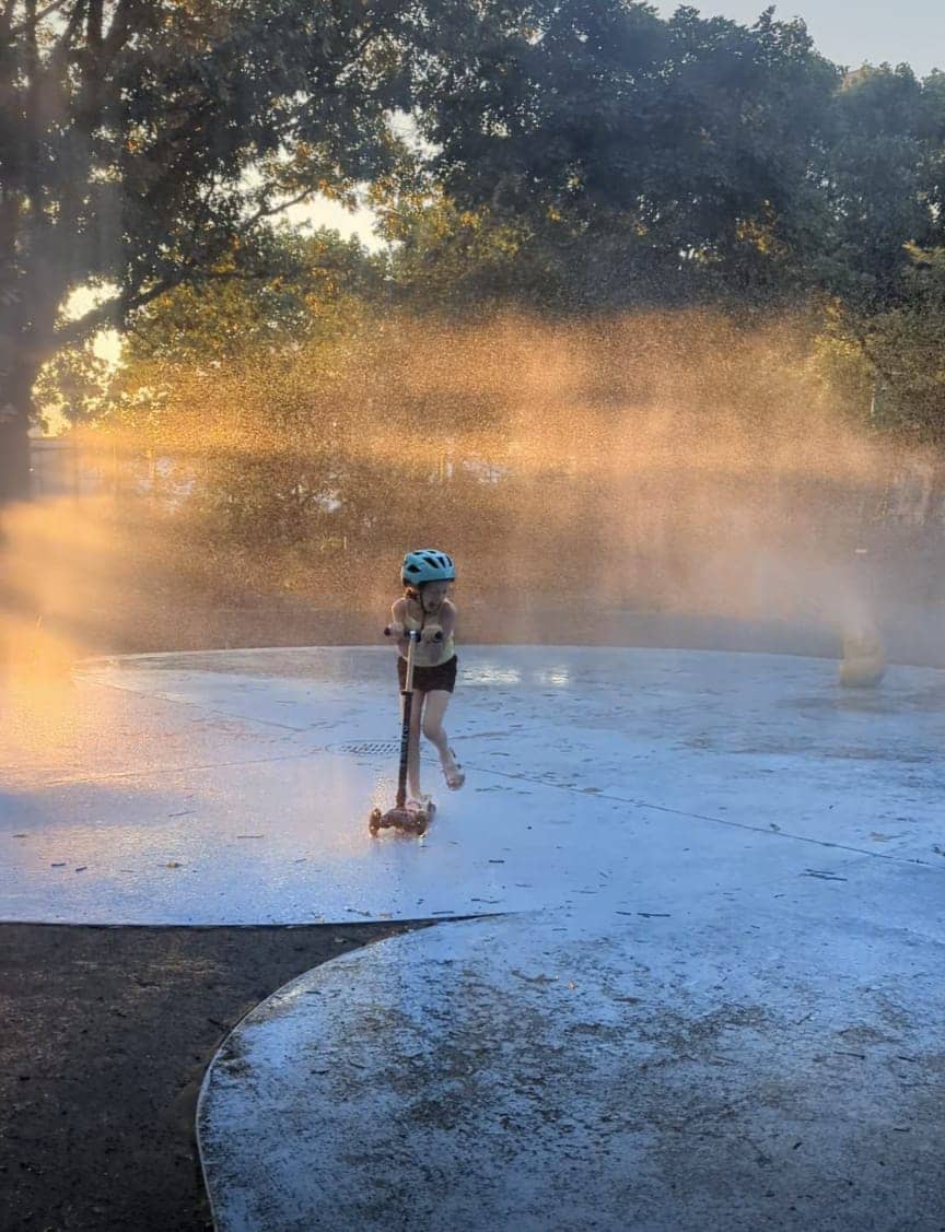 Living her best life-scootering through a sprinkler on the playground.