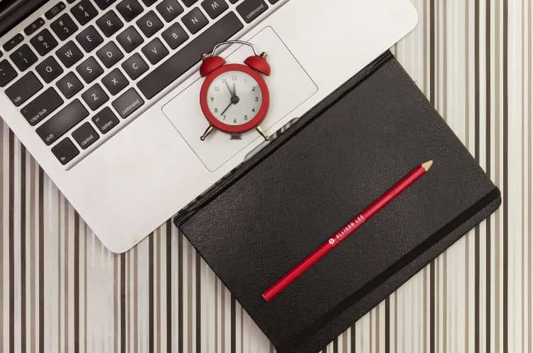 Red alarm clock on laptop keyboard beside notebook and pencil