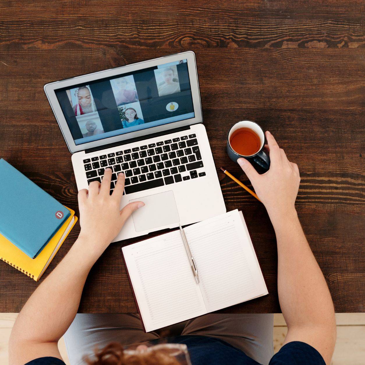 Person participating in a video meeting on a laptop with a notebook and coffee on the desk.