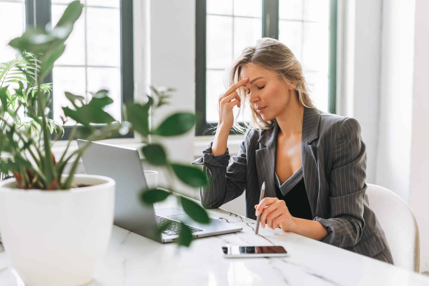 A businesswoman feels stressed while working in her office. She sits at her desk in front of her laptop, unaware that stress can age her.