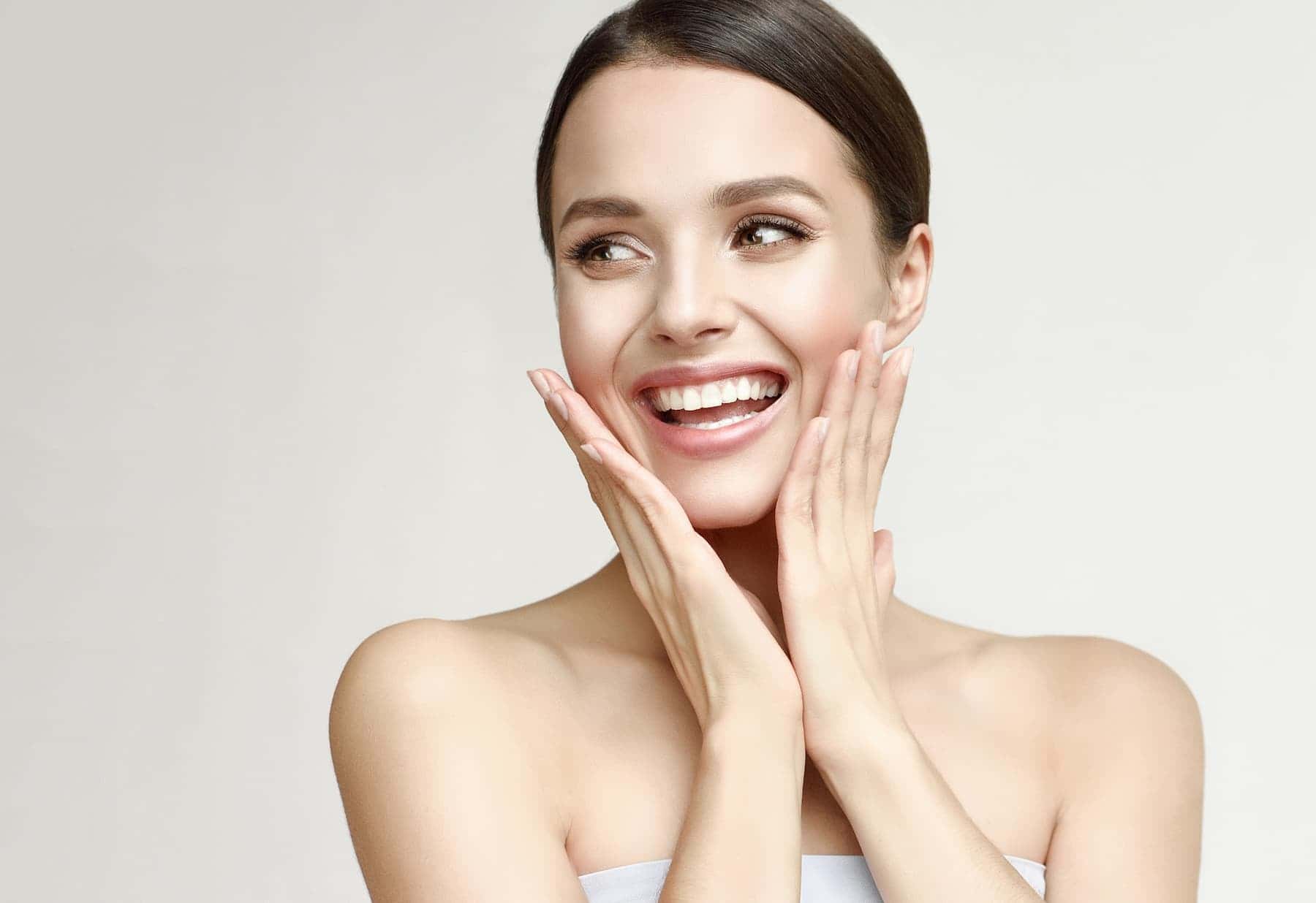 A woman smiles and touches her face against a light background, happy with the great results from using a LED mask from CurrentBody.