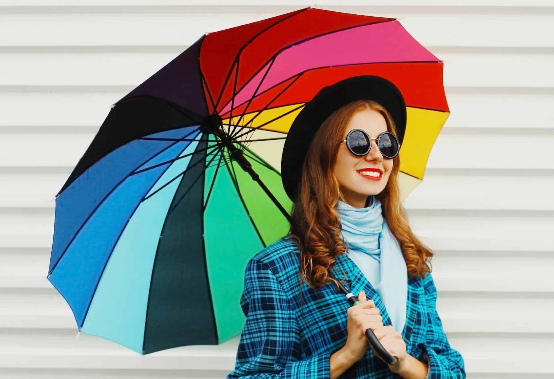A woman wearing sunglasses holds a multicolored umbrella outdoors, illustrating color psychology in wardrobe choices.