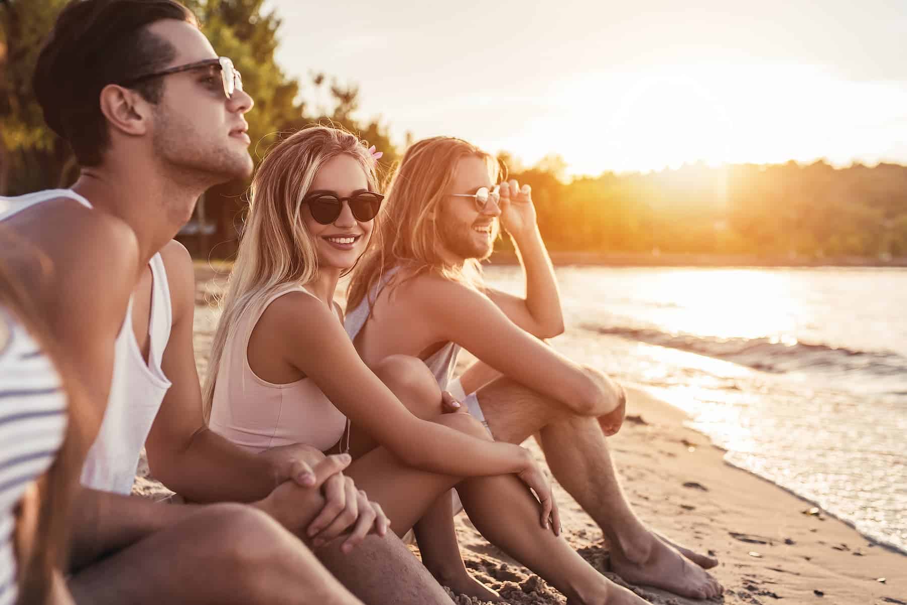 A group of young people in sunglasses relax on the beach at sunset, showing how confidence and social connection play a role in how to be attractive.