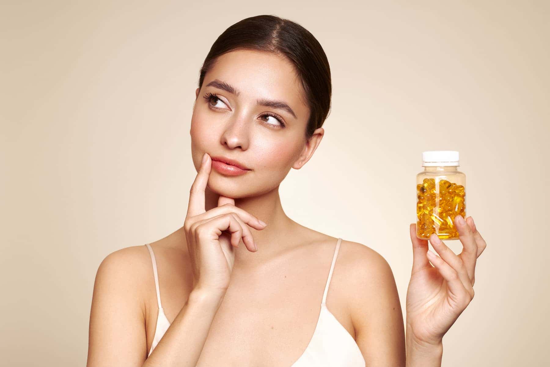 A beautiful woman holds collagen supplements in one hand, thoughtfully wondering if they truly improve her skin.