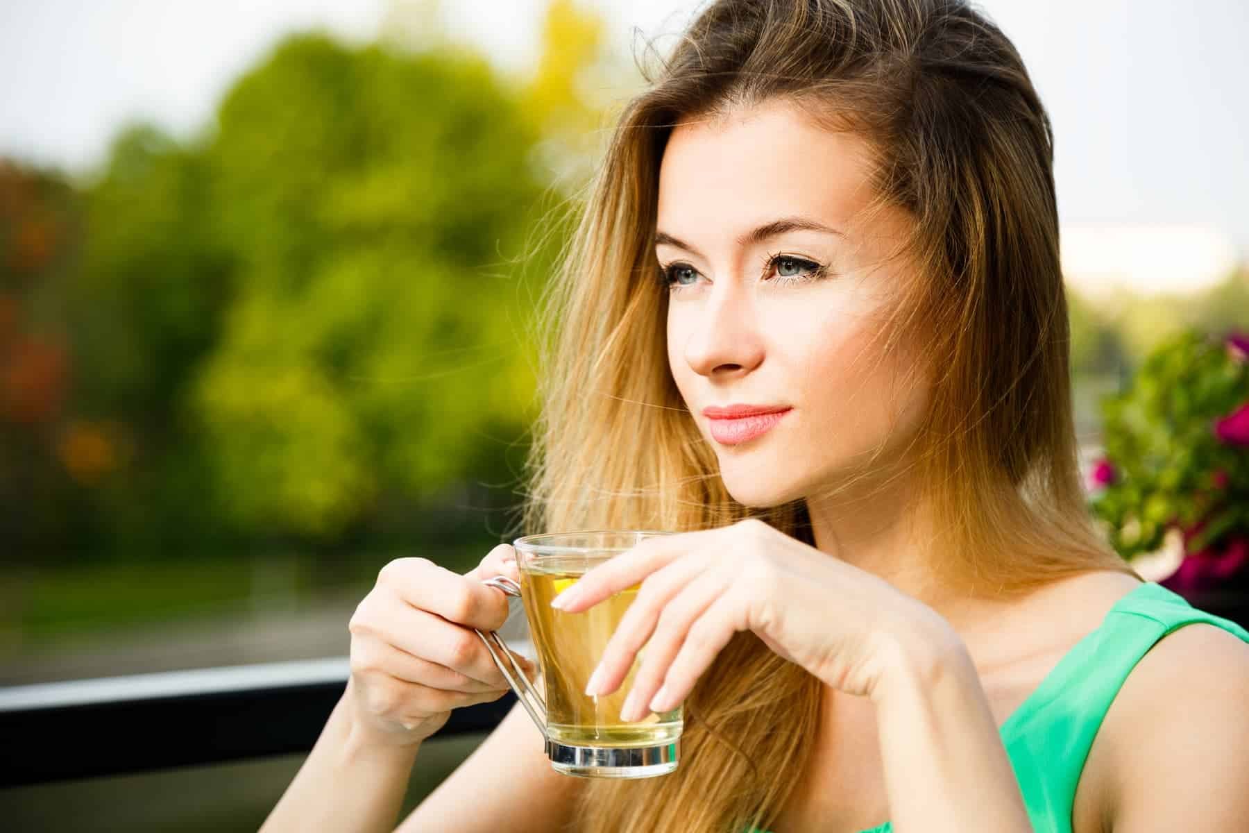 A woman drinks green tea against a lush green backdrop, showing knowledge of what are antioxidants for skin and a commitment to a healthy lifestyle.