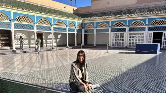 A person is sitting on a tiled floor in a courtyard featuring colorful geometric patterns. The building around the courtyard has an ornate design with arches, blue and yellow paint, and intricate latticework. A few people are walking in the background, and sunlight casts shadows across the area.