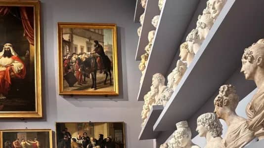A room with art exhibits, featuring several bust sculptures on shelves to the right and framed paintings on the walls. The paintings depict historical scenes and individuals. The overall atmosphere suggests a museum or gallery setting.