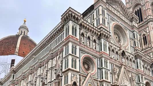 The image shows a detailed view of a large Gothic-style cathedral with intricate architectural features, including ornate carvings and circular windows. In the background, a large domed structure with a terracotta roof and an ornate golden detail at the top is partially visible. The sky is overcast.