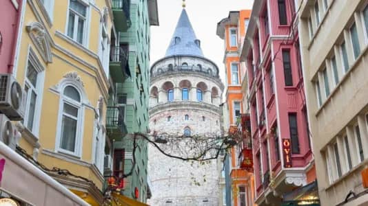 A street view leading to the Galata Tower in Istanbul. The image is framed by colorful buildings on both sides, with architectural details such as balconies and ornate window frames. The Galata Tower, an iconic stone tower with a conical roof, stands prominently in the center, its medieval structure contrasting with the vibrant facades of the surrounding buildings. The street has visible wiring and signs, capturing a lively urban atmosphere.