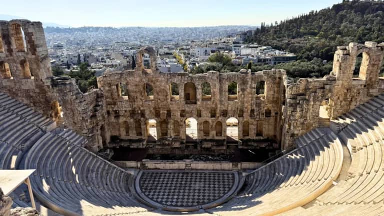 The image shows the ancient Odeon of Herodes Atticus, a large stone theater structure, with tiered seating and a semicircular stage. The front wall of the theater has arched openings. In the background, a view of Athens, Greece, is visible, with numerous buildings and trees extending to the horizon under a clear blue sky.