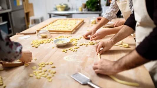 People rolling dough and shaping pasta on a wooden table, surrounded by flour and kitchen tools. A tray with formed pasta is visible in the background. The scene is in a lively kitchen setting.