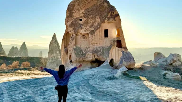 A person stands with arms outstretched, facing a large, ancient cave dwelling carved into a rock formation in Cappadocia, Turkey. The morning light casts soft shadows across the rocky landscape, with more unique rock formations visible in the background.