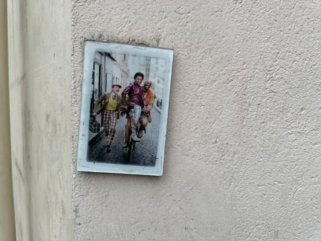 Three people share one bicycle on a cobblestone street, all caught mid-laughter — a woman in a burgundy jacket riding in the middle, a man steering behind her, and a third person in a checked suit running alongside. The photograph sits in a weathered whit