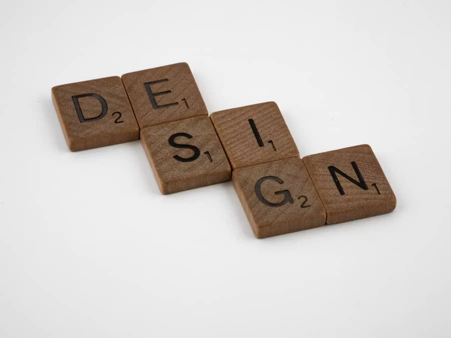 Wooden letter tiles arranged to spell “DESIGN” in a staggered, zigzag pattern on a clean white background. The tiles resemble Scrabble pieces, with engraved black letters and small point values in the bottom-right corners. The warm wood tones contrast wit