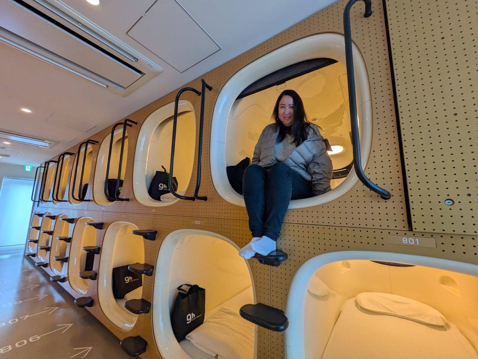 Author Malia Yoshioka sits in the top pod of a capsule hotel in Tokyo, there are two rows of pods with the openings facing out line up on a corridor.