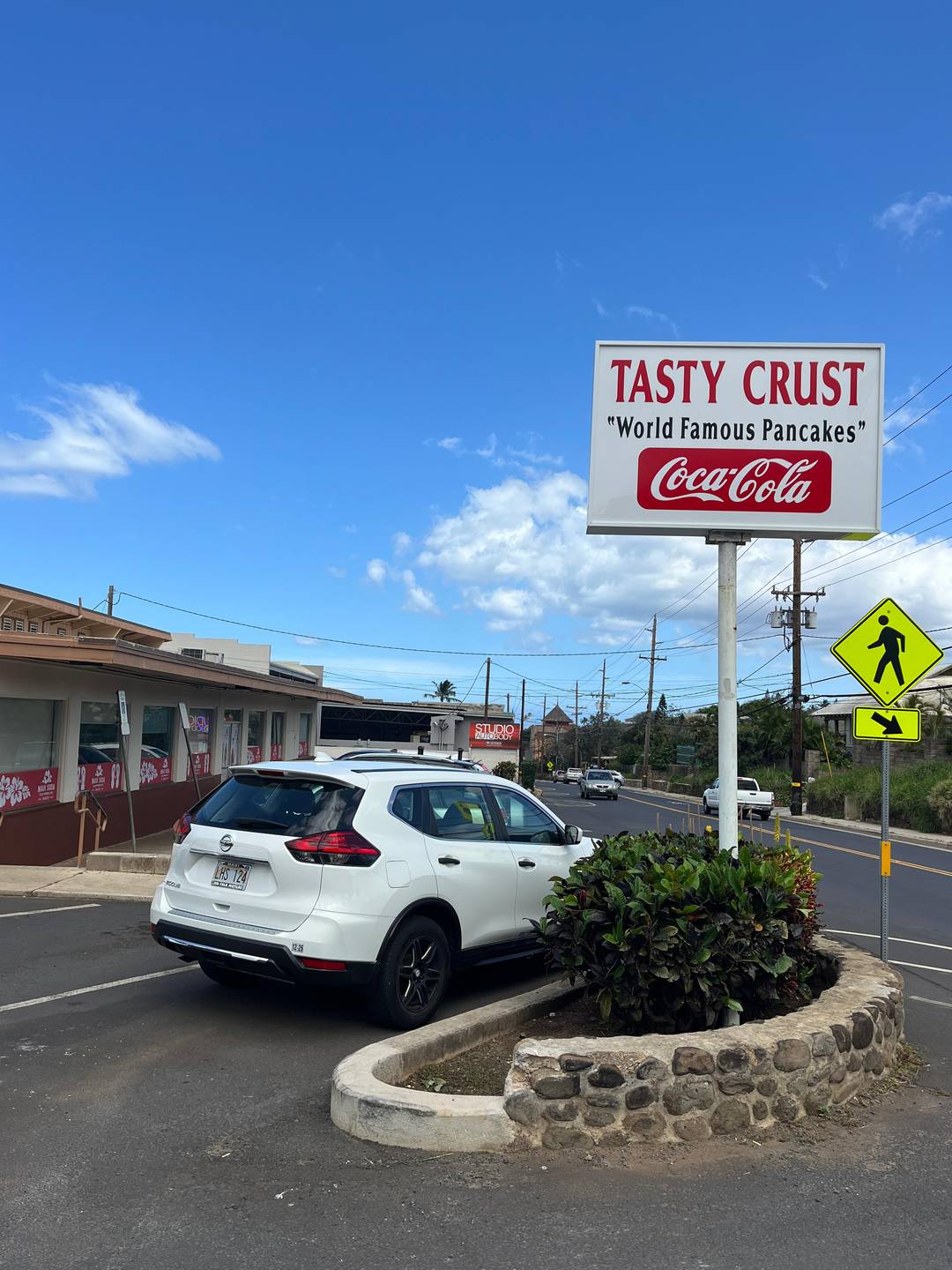 The sign outside of Tasty Crust in Wailuku Maui, reads "Tasty Crust" World Famous Pancakes" above a red and white Coca-Cola logo. The local diner is visible with its parking lot to the left, a Wailuku road to the right and blue skies above.