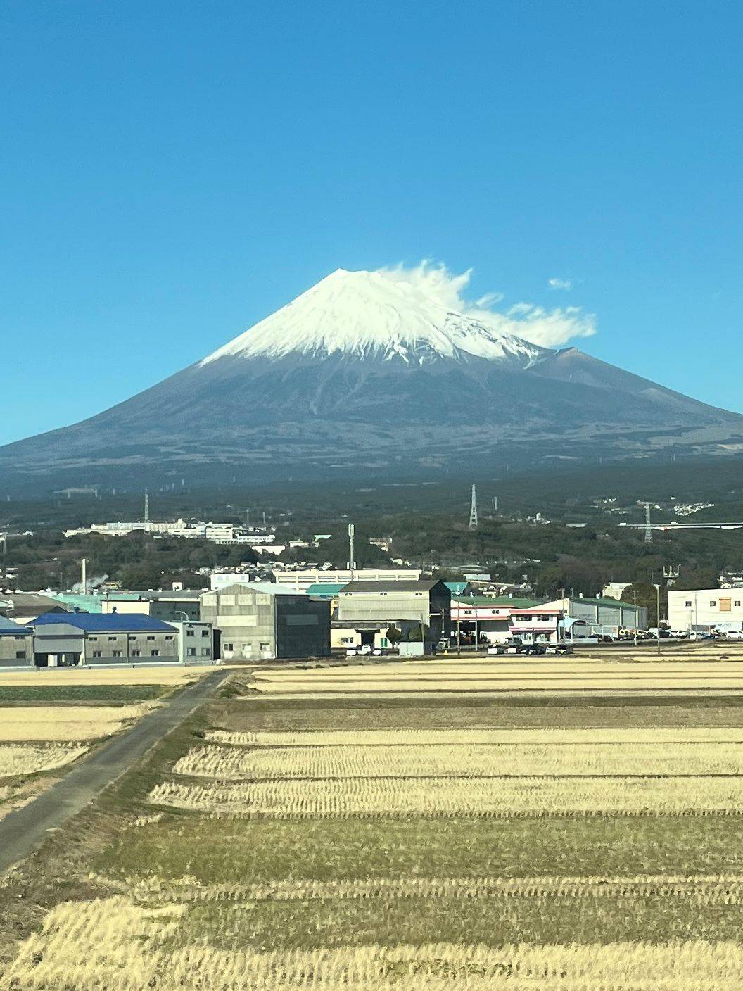 A clear view of Mt Fuji in Japan with blue sky and a white snowcap, blowing off the right side in the wind, with fields and a small town buildings in front.