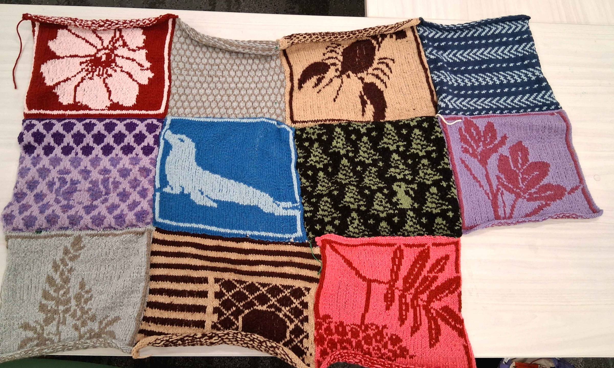 A quilt made of handknit squares is in the process of being sewen together. The squares are each knit in two colors, depicting a variety of plants and animals.
