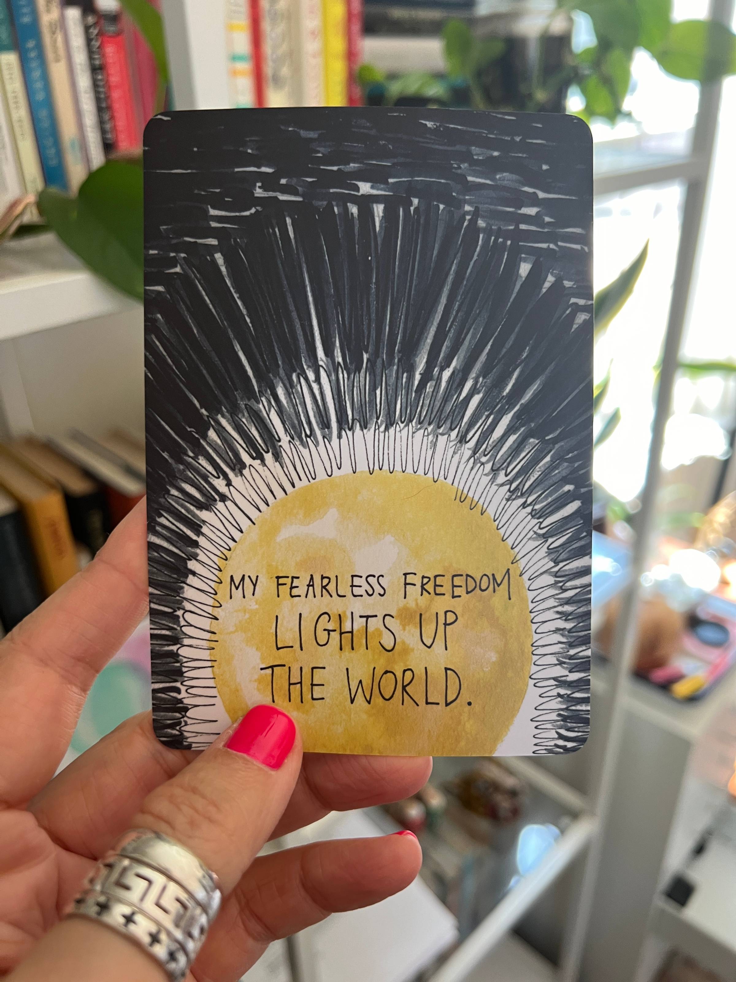 A woman’s hand with pink nail polish holding a card that has the image of a bright sun with beams lighting up the darkness and the words “My fearless freedom lights up the world.”