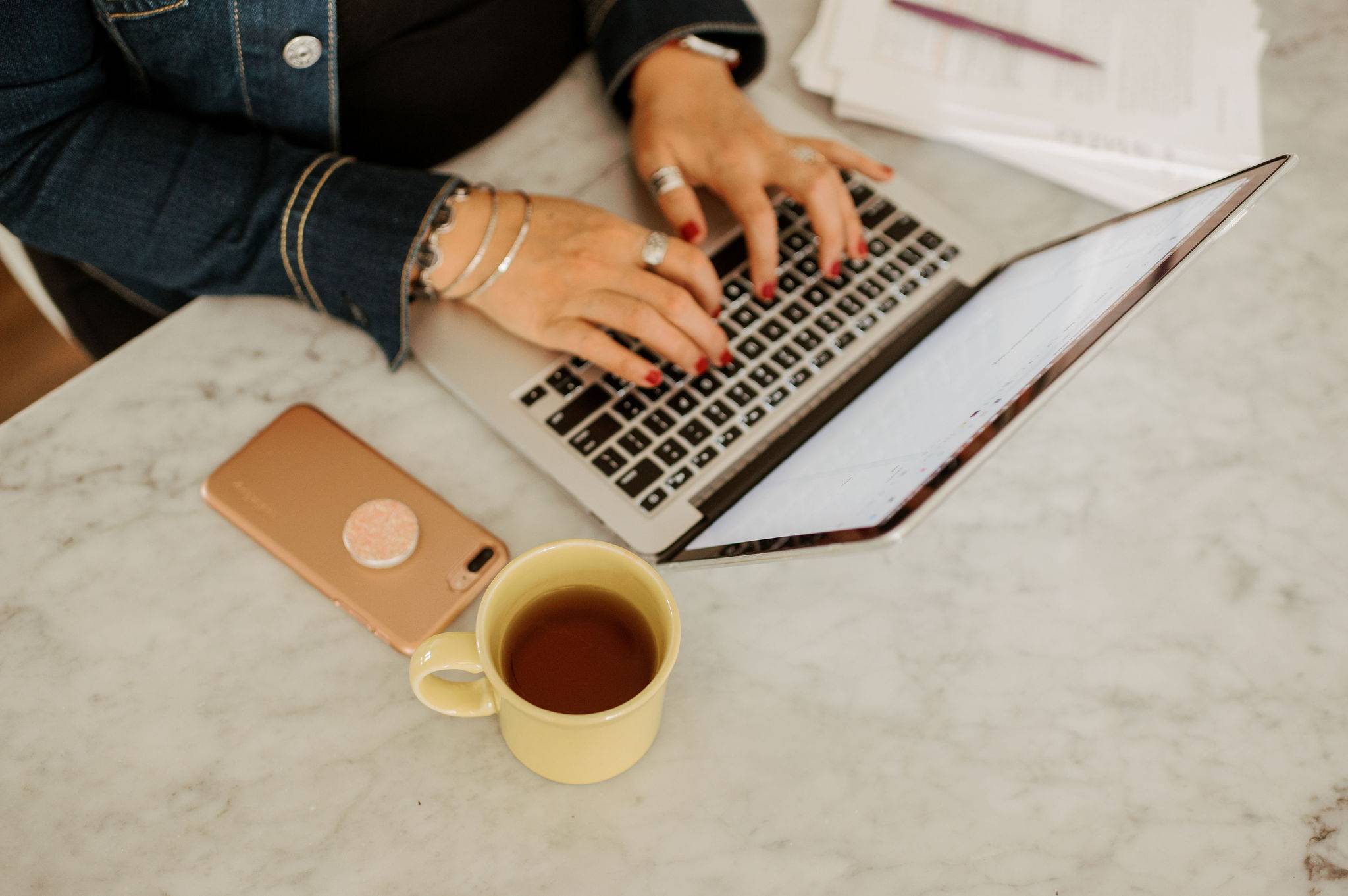 A woman's hands typing on her laptop computer, with a stack of papers next to her on one side and her phone and a coffee mug on the other side.