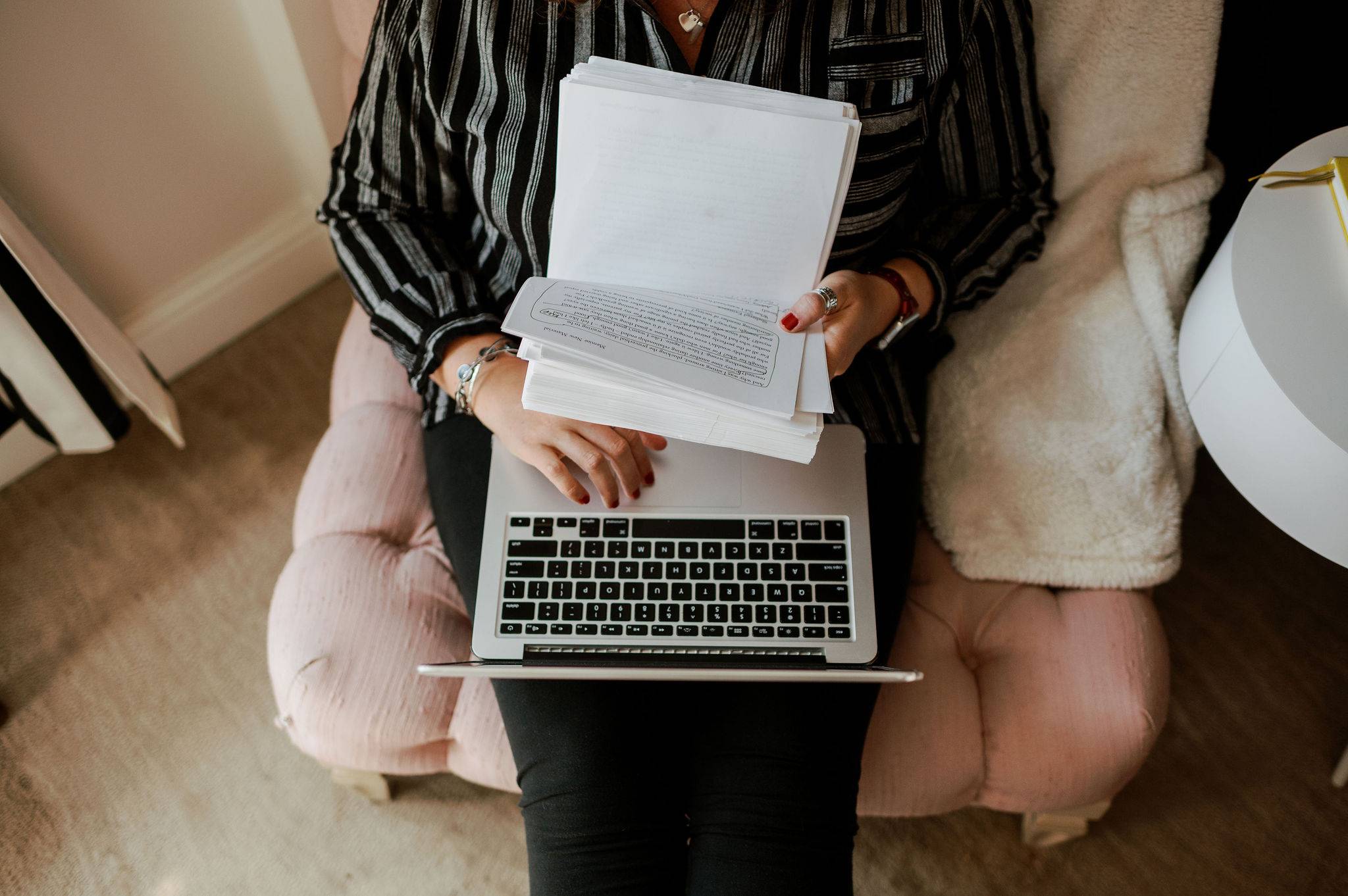 A woman sitting in a comfortable chair with a laptop and printed manuscript pages on her lap.