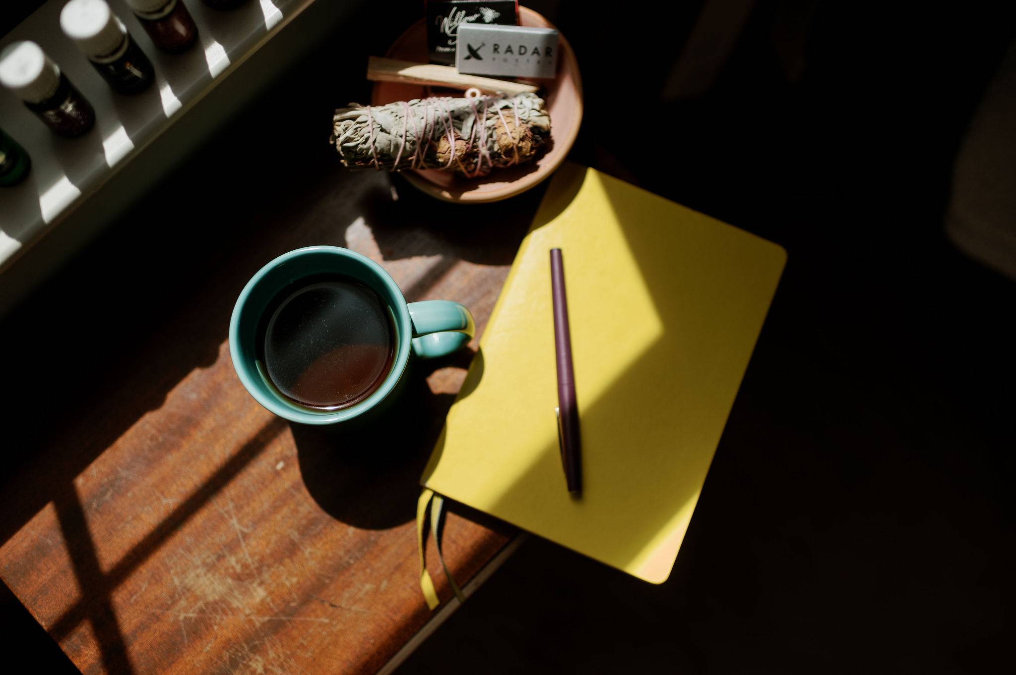 A turquoise coffee mug of tea or coffee next to a yellow journal with a purple pen resting on it. Light from the window makes a shadow across the photo.