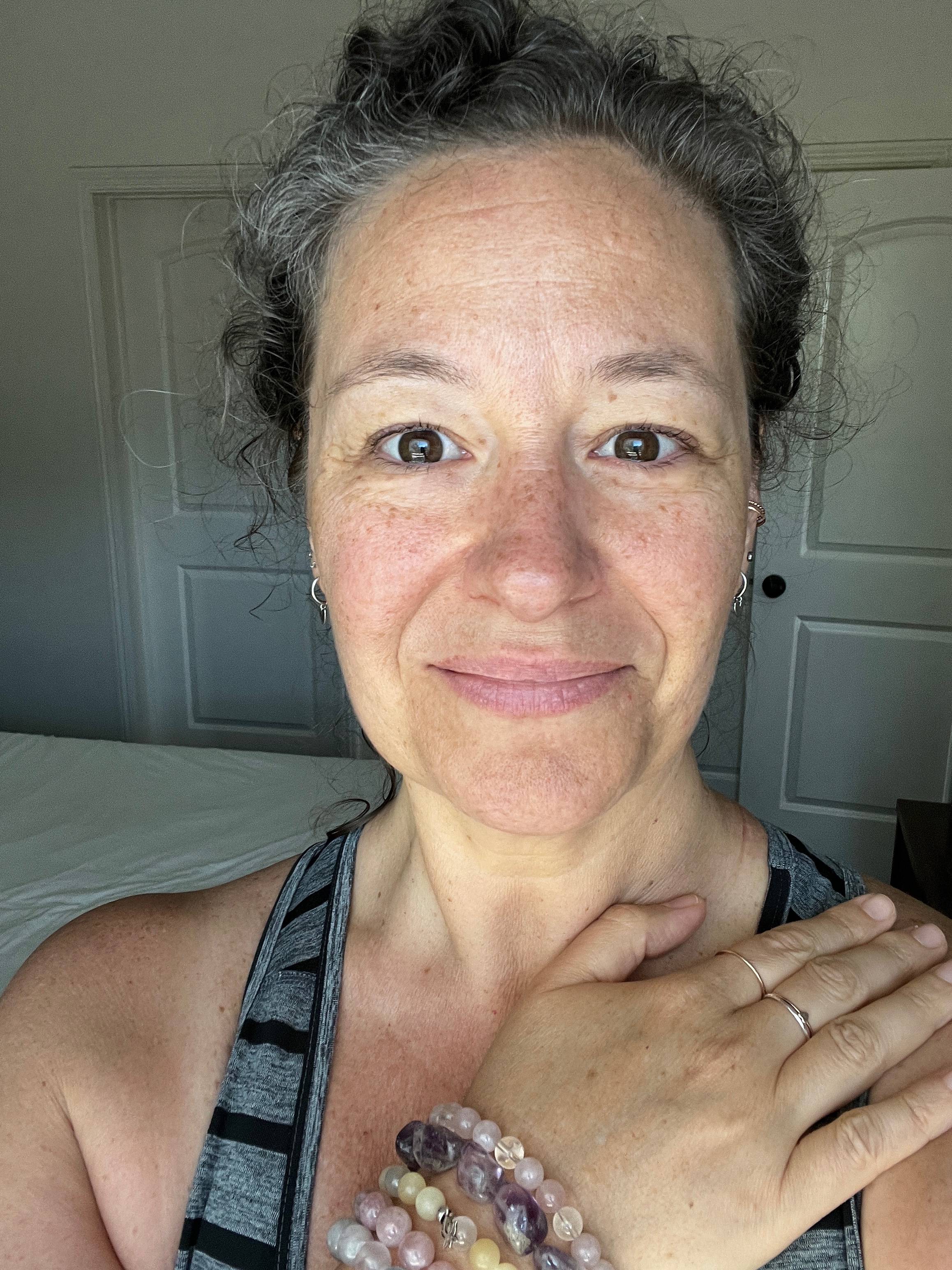 Close up of a woman's face with a slight smile. Her hand is over her heart and she wears gemstone bracelets.