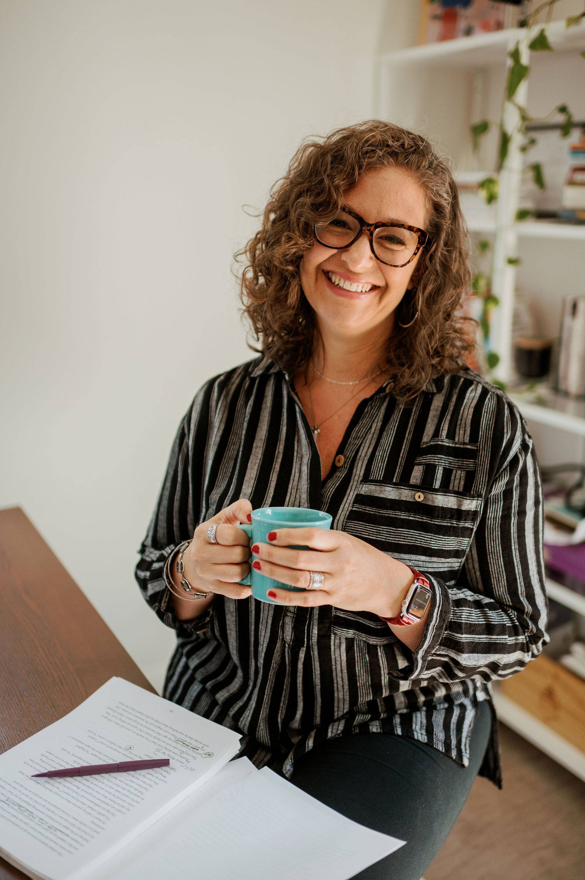 Woman with curly hair, wearing glasses, sitting at a desk with a manuscript and pen resting in front of her. She's holding a coffee mug and smiling. 