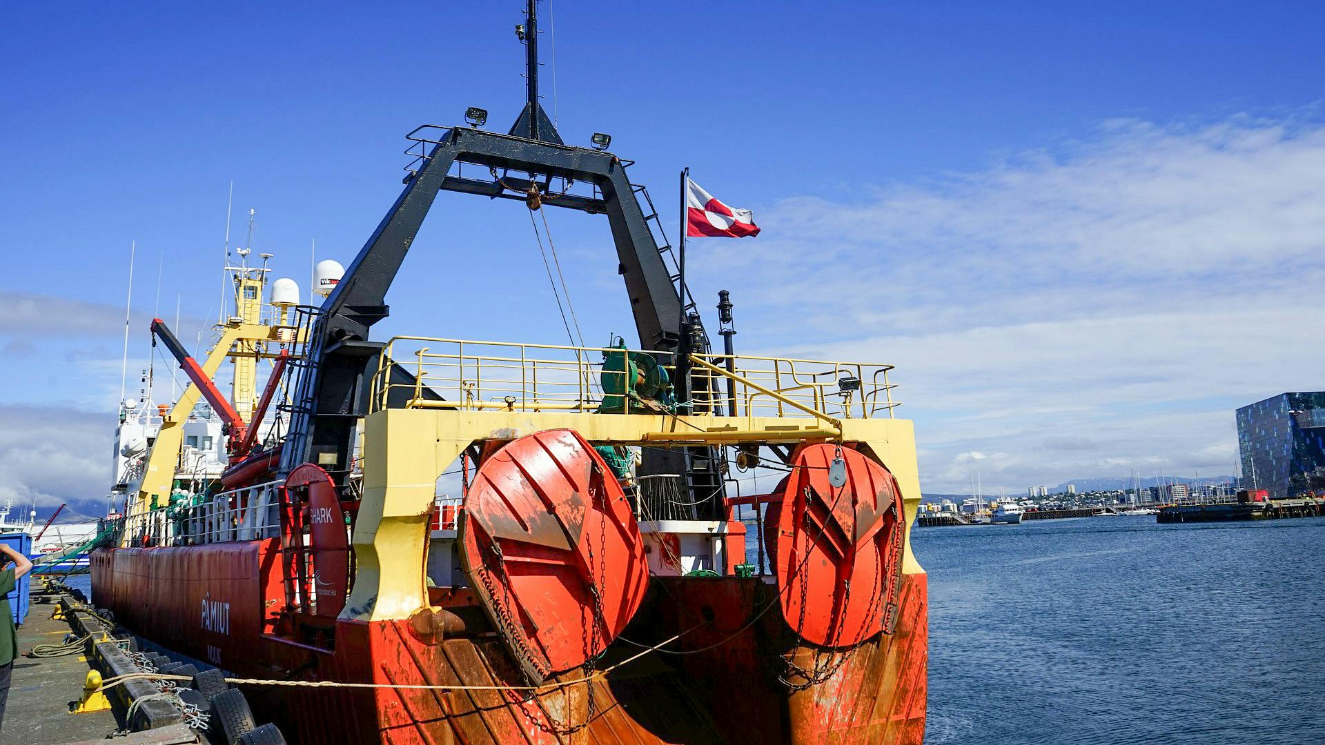 Commercial whaling vessel docked at a harbor, showing large red circular harpoon reels and a yellow A-frame structure mounted on deck. The ship flies a Greenlandic flag and has industrial whaling equipment visible. The harbor scene includes a modern city 