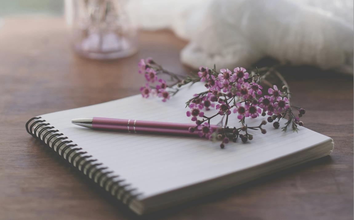 Open journal with pink pen and soft pink flowers on a wooden table by a window, representing grief journal prompts for losing a parent.